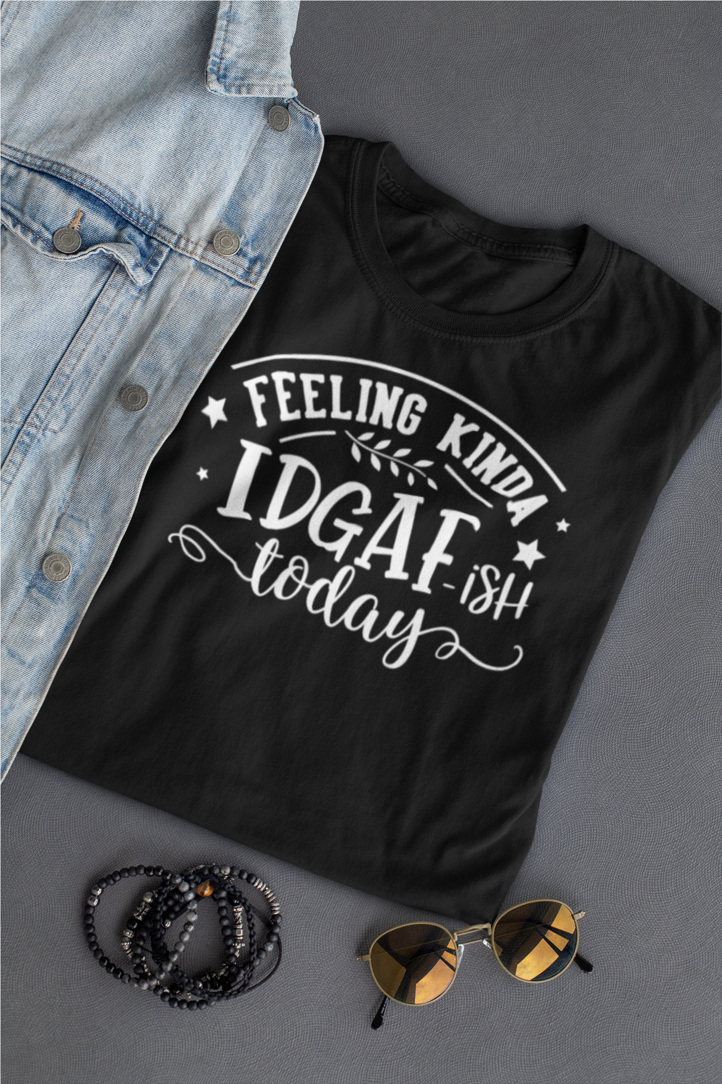 A black folded t shirt with white writing that says Feeling Kinda IDGAF ish today laying next to a jean jacket, sunglasses and bracelets
