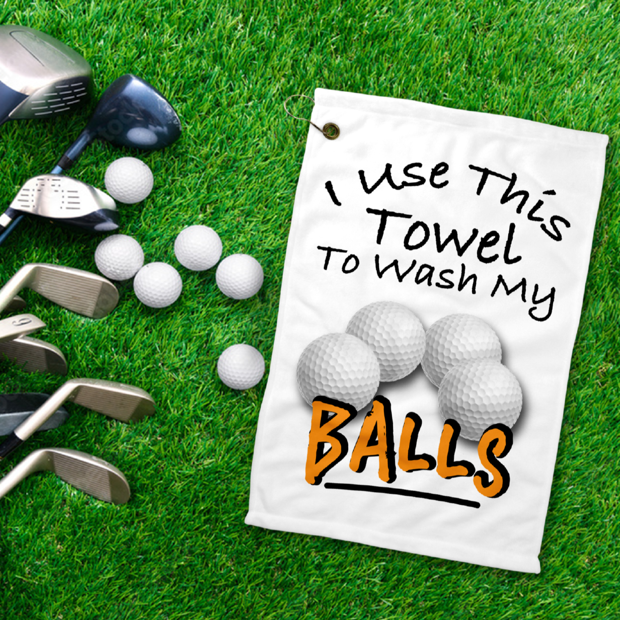 A 12x18 inch white golf towel laying on the grass next to golf clubs, that says in black writing Use this towel to wash my balls. With a picture of four golf balls