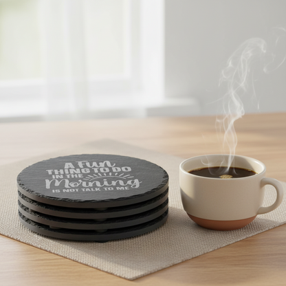 A round black slate coaster with laser etched text that reads 'A Fun THING TO DO IN THE Morning IS NOT TALK TO ME' with a small sun graphic, shown on a morning coffee table with a cup of black coffee.