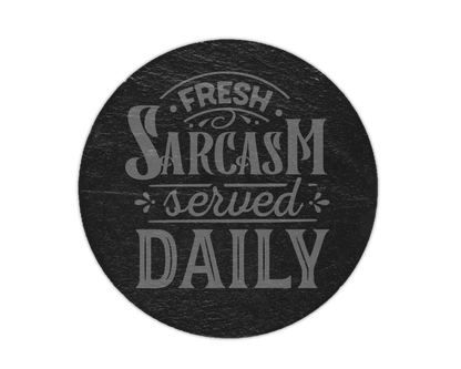A round black slate coaster or plaque featuring the phrase 'Fresh Sarcasm served DAILY' in a light gray stylized font, shown against a plain white background.