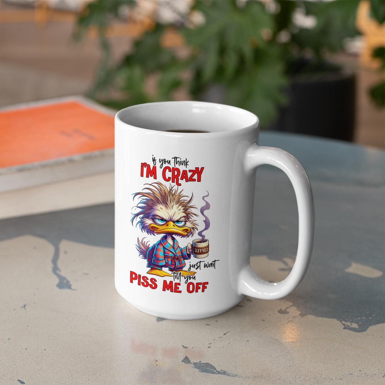 A white coffee mug sitting on a out door table with a cartoon male duck in pajamas holding a coffee mug and text that says i you think I'm crazy just wait till you piss me off