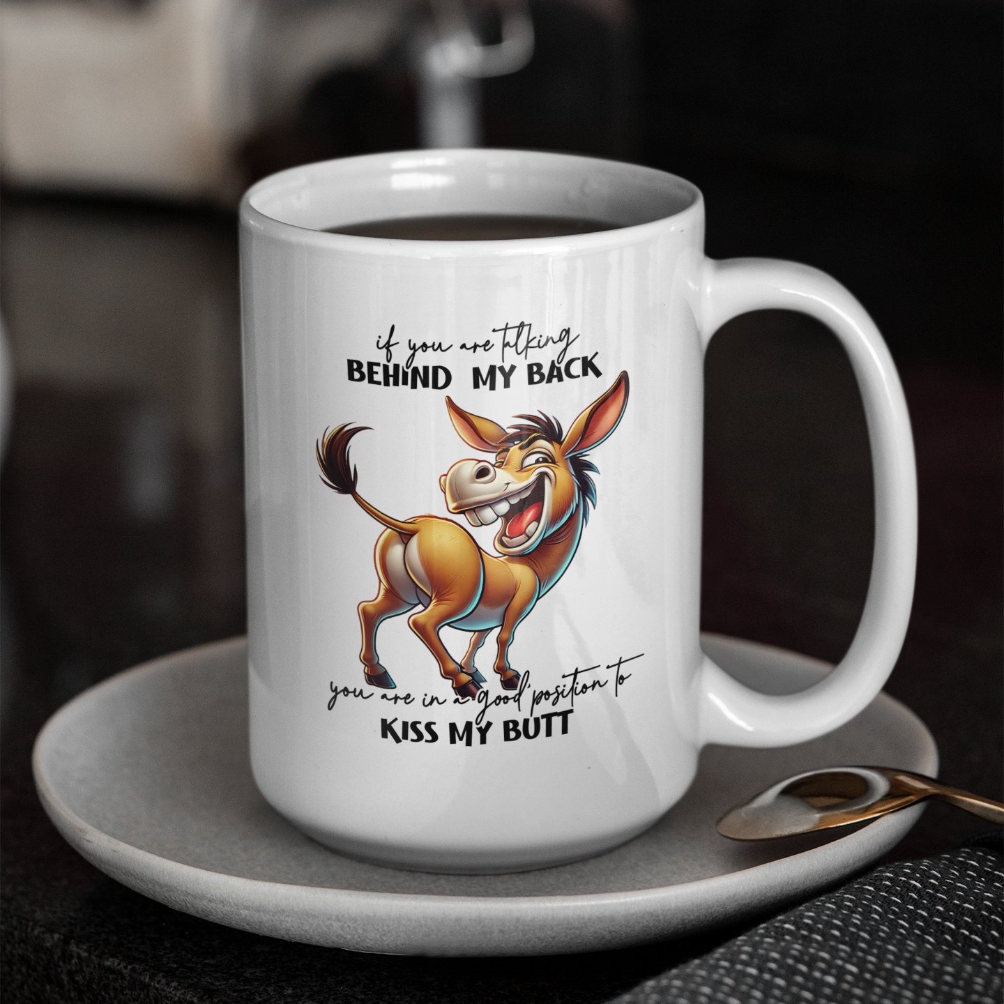 A white coffee mug  on a saucer with a donkey on it that says If you are talking behind my back you are in a good position to kiss my butt