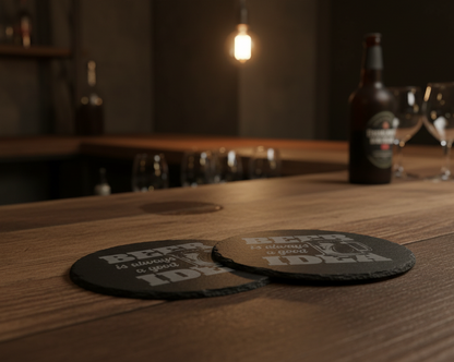 Two round black slate coasters shown on a white background, with one front-facing and one back-facing. The front coaster has a gray laser etched design reading “BEER is always a good IDEA” with a beer mug illustration.
