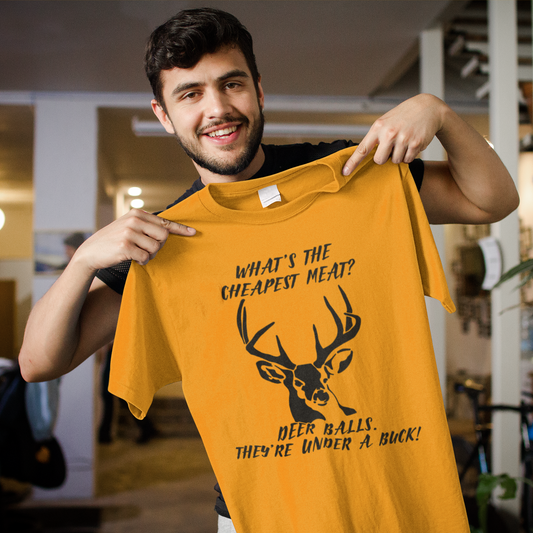 A young man hold up an orange t-shirt with a deer on the front chest that says what's the cheapest meat? deer balls. they're under a buck!